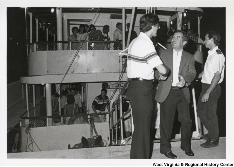 Representative Nick J. Rahall (D-W.Va.) shakes hands with an unidentified man in front of a large group of unidentified people on a ship.